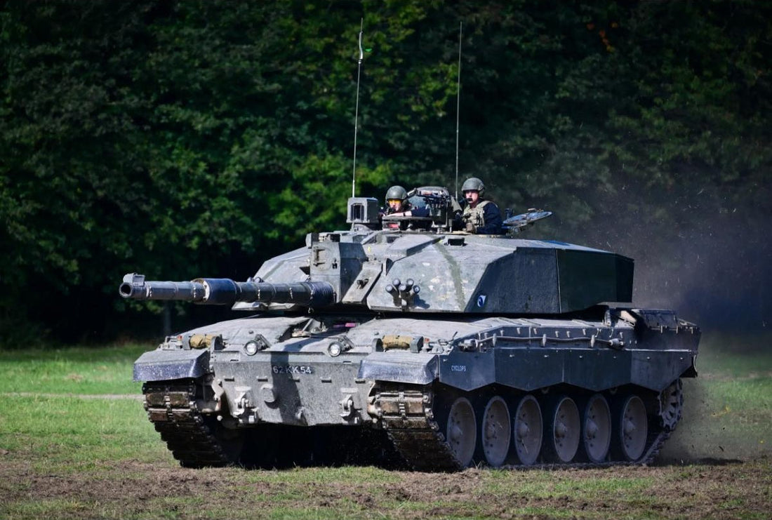 ritish Challenger 2 Main Battle Tank moving across a grassy field during a military exercise, with two crew members visible in the turret.
