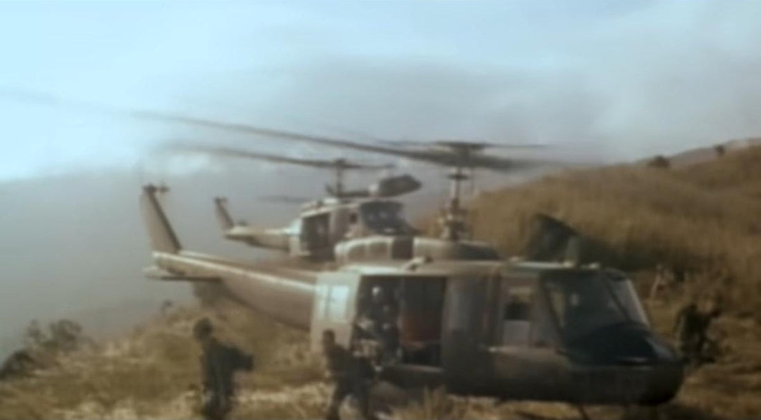 Two UH-1 "Huey" military helicopters landing on a dry, grassy, and hazy hillside. The side door of the main helicopter is open, with soldiers visible inside. The spinning rotors are kicking up dust and haze.