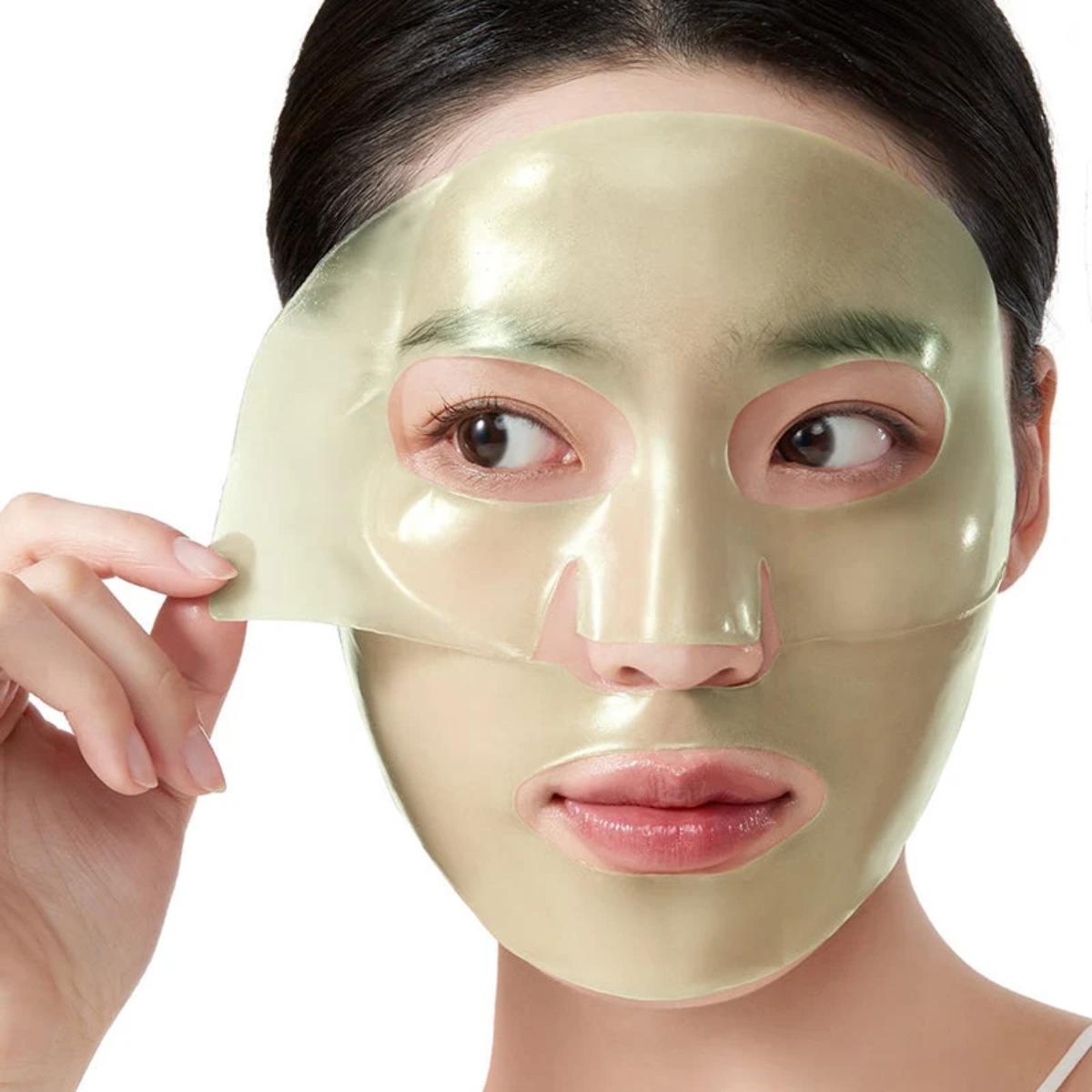 Close-up of an Asian woman's face with a glossy, light-green Anua Heartleaf 70 Soothing Collagen hydrogel sheet mask partially peeled off, isolated on a white background