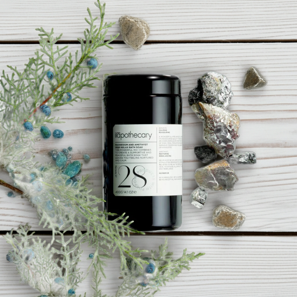 ila Apothecary product displayed on a white wooden surface with decorative stones and green flowering branches, styled in a clean natural wellness setting.