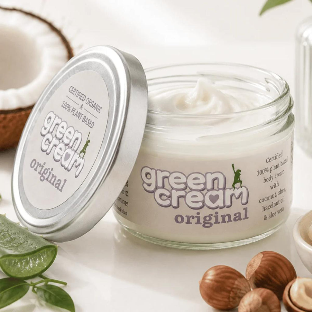 The Green Woman product - A jar of Green Cream Original plant-based body cream with its lid leaning against it, styled with coconut, aloe vera, and nutmegs on a bright white surface.