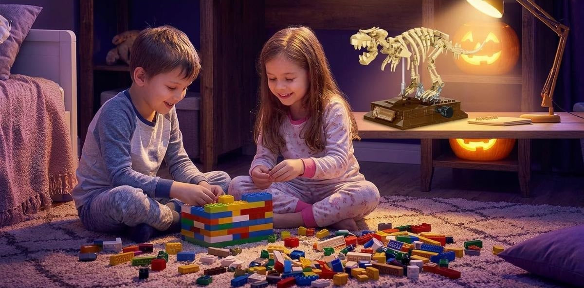 A cozy children's bedroom in deep purple and golden Halloween lighting, with a boy and a girl playing with building bricks on the carpet, a dinosaur skeleton block model on the desk, and glowing jack-o'-lantern pumpkins in the background.