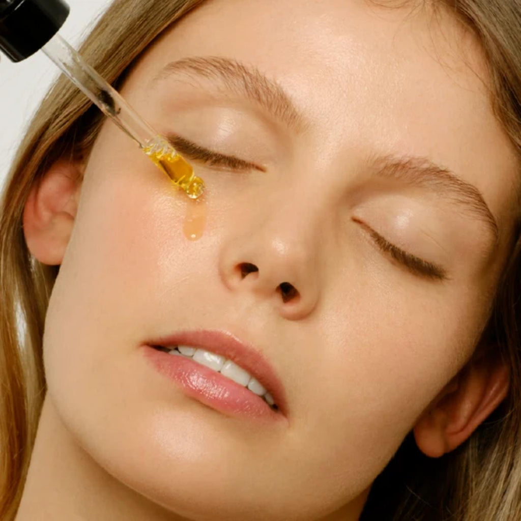 A close-up of a woman with her eyes closed as a golden amber facial serum is being dropper-fed onto her face.