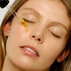 A close-up of a woman with her eyes closed as a golden amber facial serum is being dropper-fed onto her face.