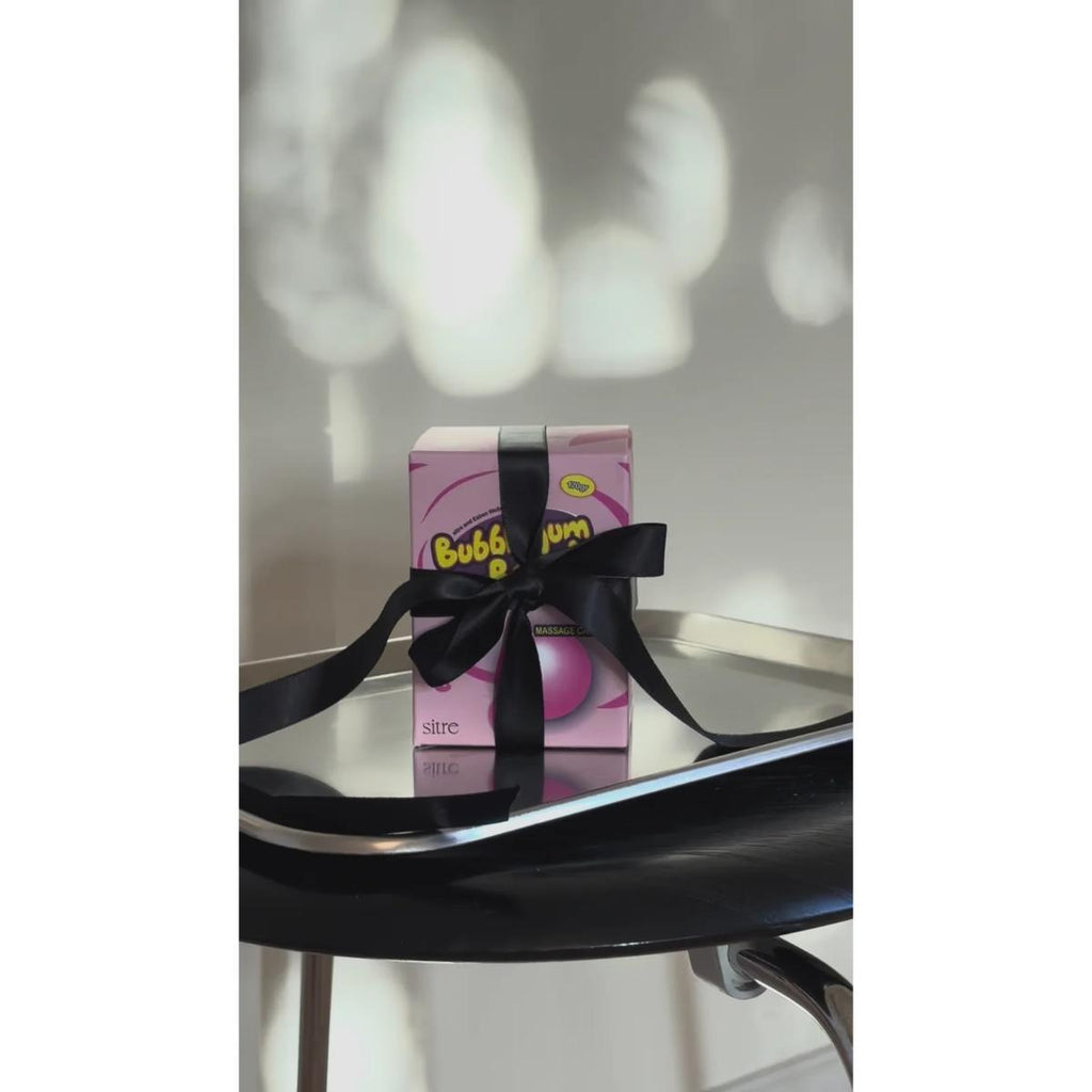 A pink Bubble Gum gift box tied with a black ribbon, displayed on a reflective black table against a light wall with soft shadow patterns.