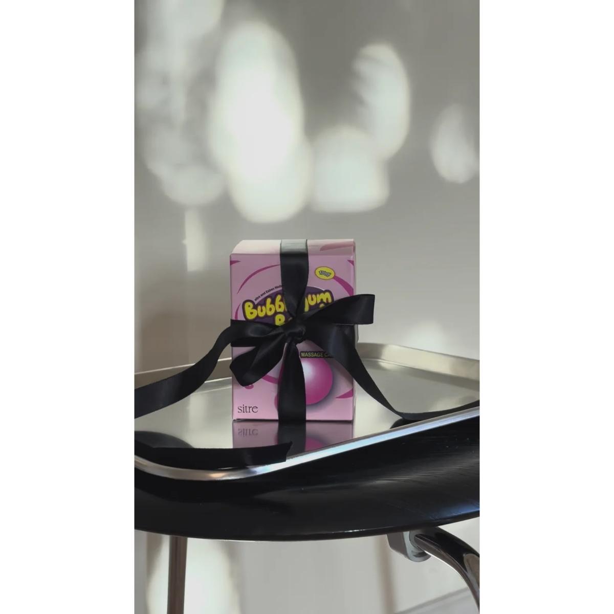 A pink Bubble Gum gift box tied with a black ribbon, displayed on a reflective black table against a light wall with soft shadow patterns.